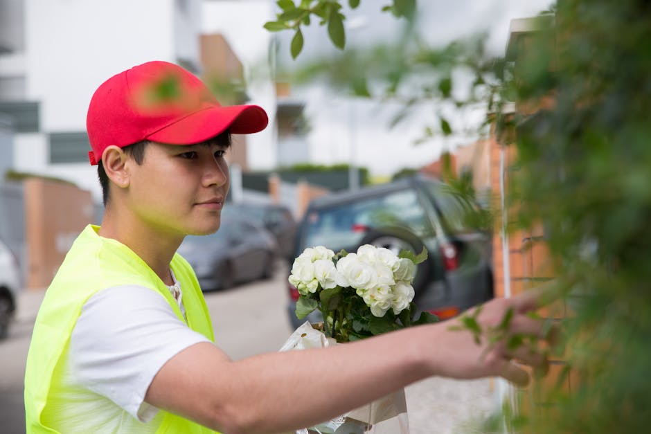 A delivery person, wearing a bright yellow high-visibility vest and a red cap, is seen handing over a bouquet of fresh white flowers, likely hydrangeas or similar blooms, wrapped in white paper, to a
