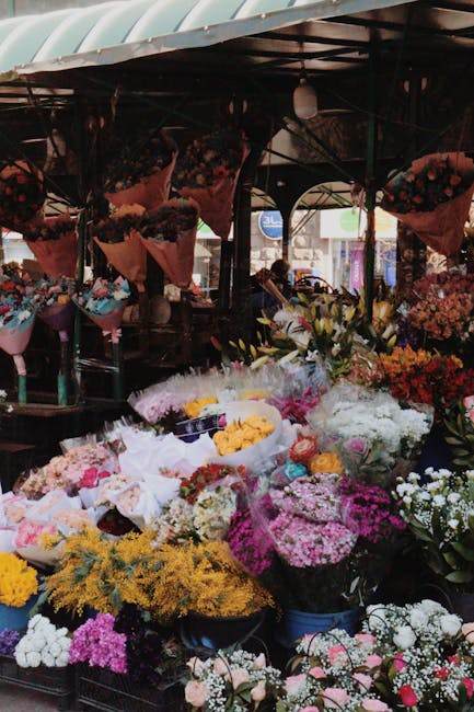 A vibrant display of fresh flowers at an outdoor market stall, arranged in various bouquets and floral arrangements. The flowers include bright yellow, pink, white, purple, and red blooms, with some w