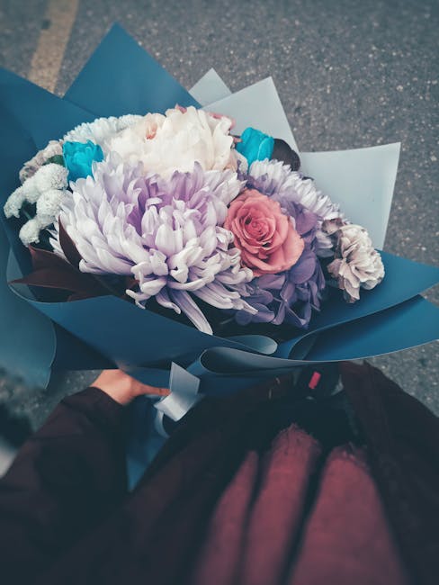 A vibrant bouquet of fresh flowers featuring a large, pale purple chrysanthemum, soft pink roses, white and purple flowers, accented with small bright turquoise blooms. The arrangement is wrapped in m