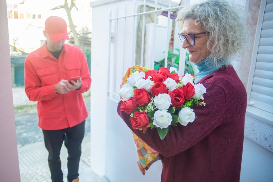 A woman with curly gray hair and glasses, dressed in a maroon cardigan and blue turtleneck, holding a vibrant bouquet of fresh red and white roses wrapped in yellow patterned paper, standing near a wh