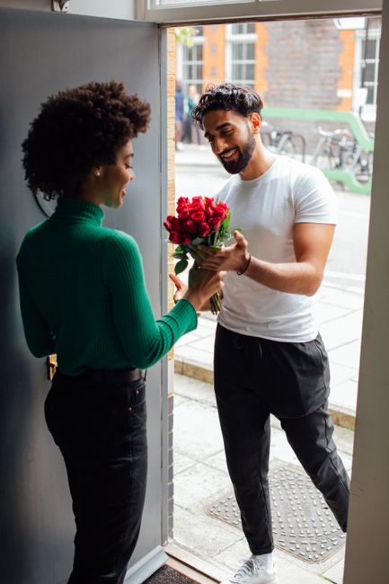 A man with dark, curly hair and a beard, wearing a white t-shirt and black trousers, is smiling as he hands a bouquet of vibrant red roses to a woman with an afro hairstyle, dressed in a green, long-s